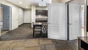 A modern kitchen with dark cabinetry, a dining area with a patterned tablecloth, and a hallway with a carpeted floor. - Homewood Suites by Hilton Waco (Waco)