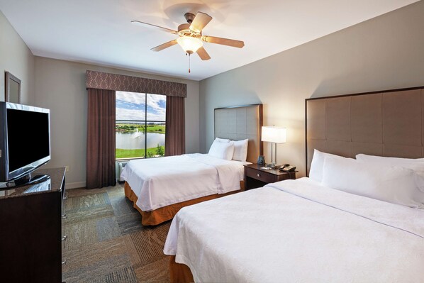 A hotel room with two beds, a television, a ceiling fan, a window with a view of greenery, and a nightstand with a lamp.