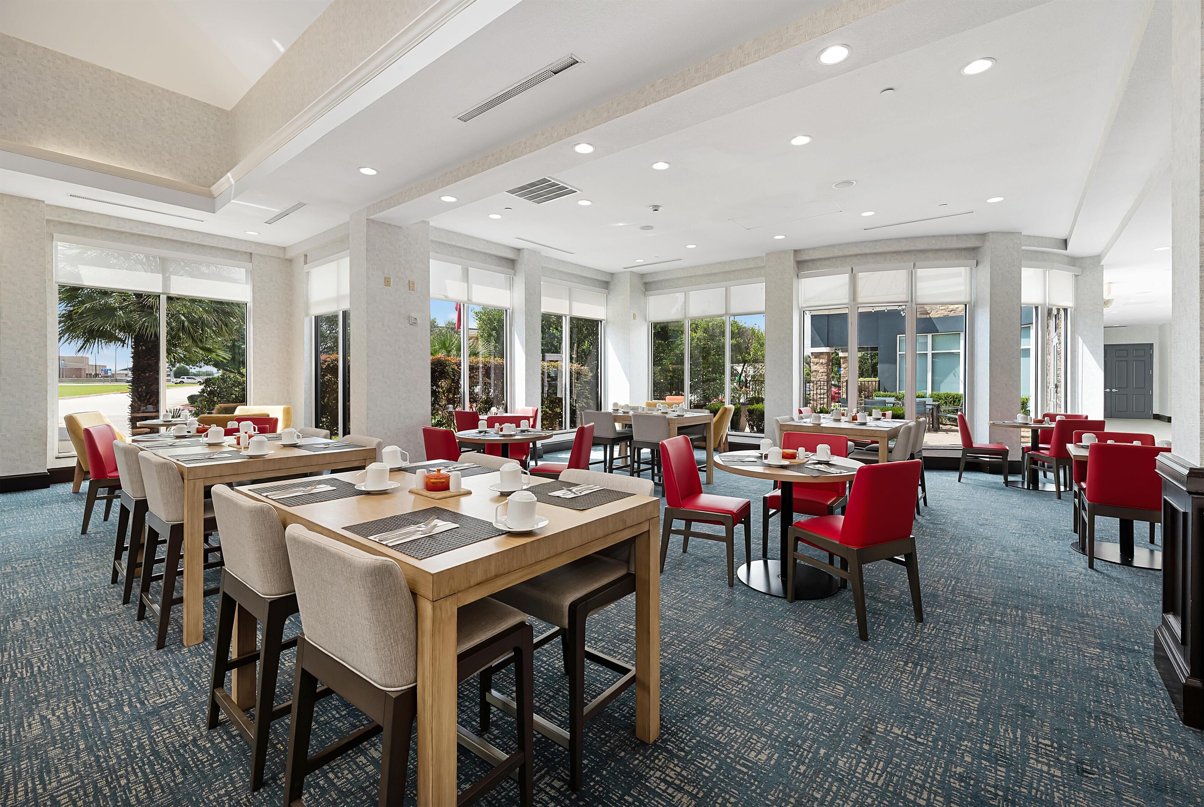 Un vaste espace dédié à la salle à manger, avec de nombreuses tables et chaises, de grandes fenêtres donnant sur l’extérieur et un sol recouvert de moquette.