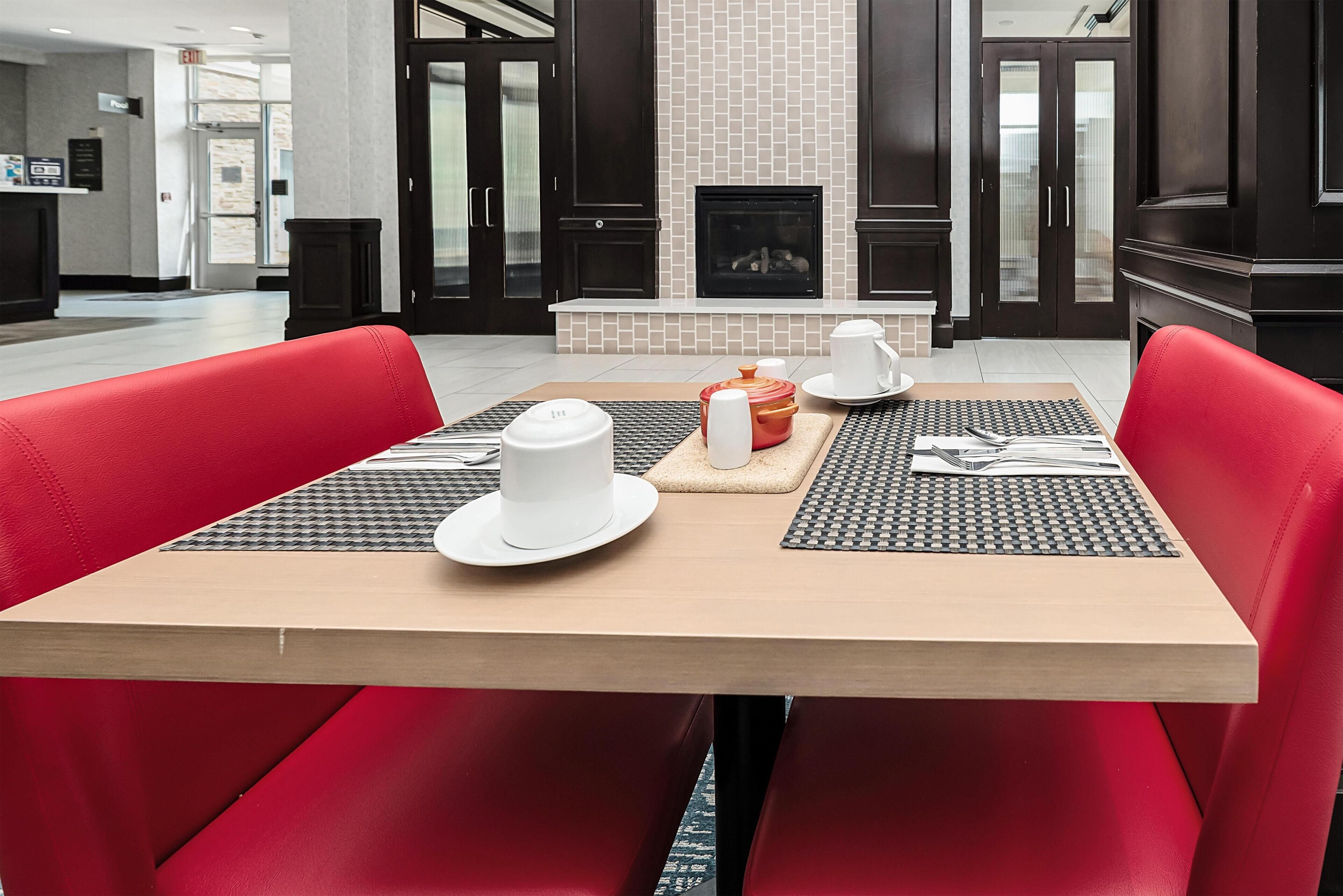 Een eethoek met een tafel gedekt voor twee personen, rode bekleden stoelen, een open haard en een receptiebalie op de achtergrond.