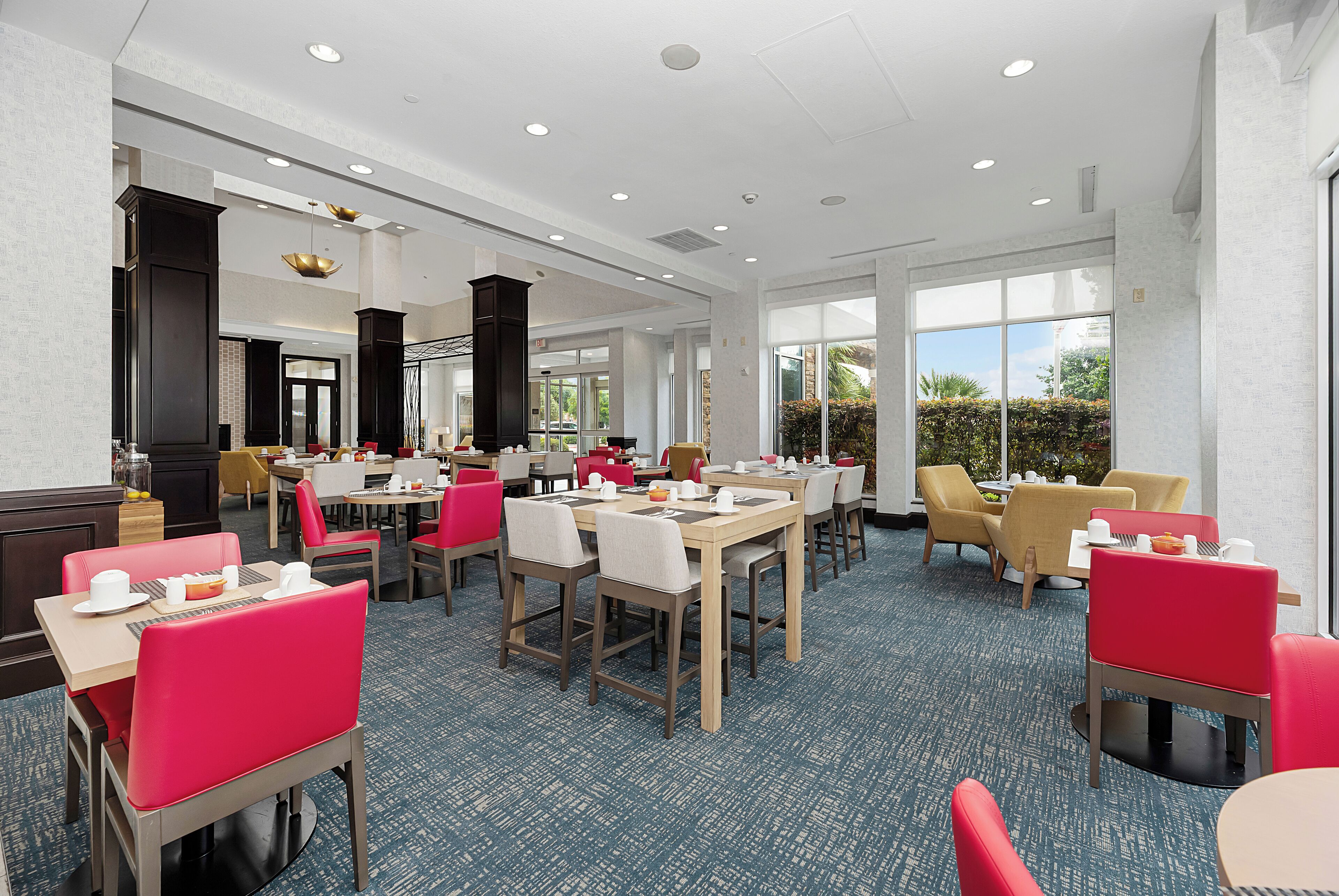 A spacious dining area with multiple tables and chairs, large windows offering views of the outdoors, and a modern interior design.