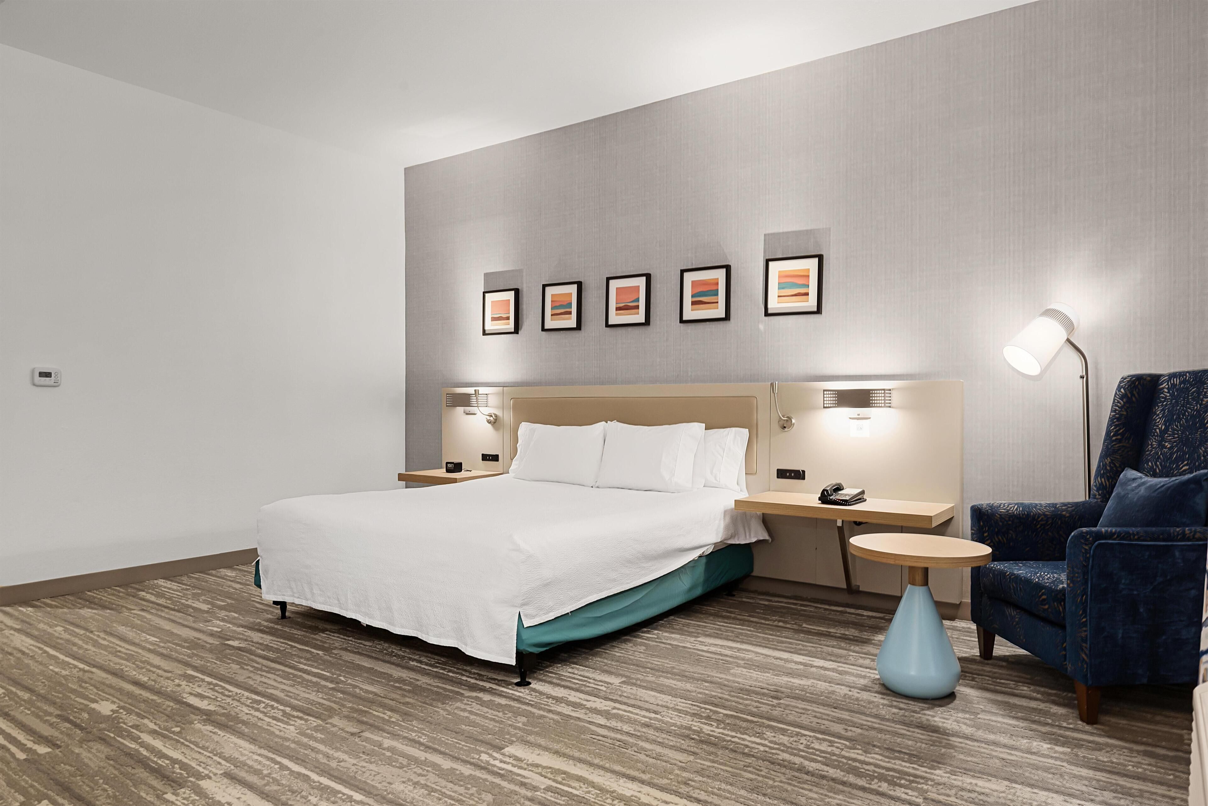 A hotel room with a bed, a desk, a chair, a lamp, and framed pictures on the wall.