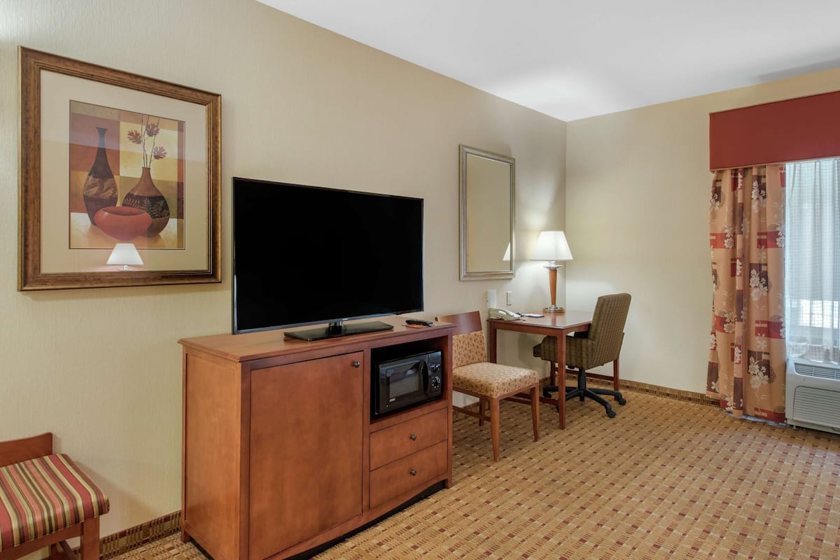 Quarto de hotel com televisão de ecrã plano, micro-ondas, área de refeições com mesa e cadeiras, candeeiro e uma obra de arte emoldurada na parede.