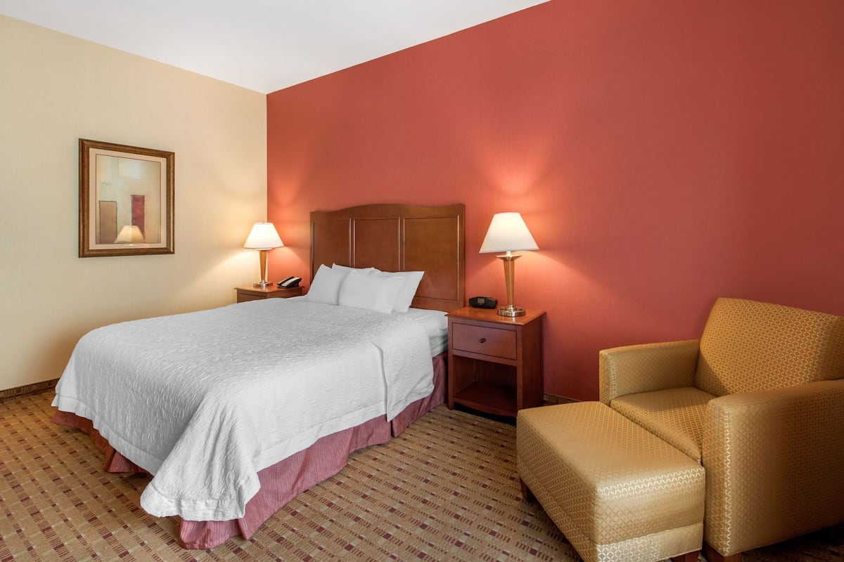Quarto de hotel com uma cama, duas mesas de cabeceira com luminárias, uma cadeira e um quadro na parede.