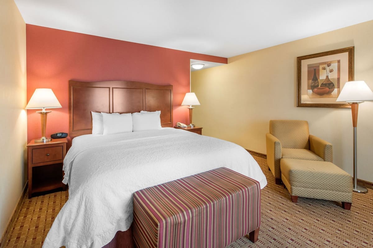 Quarto de hotel com uma cama grande, duas luminárias de cabeceira, uma cadeira e uma mesinha.