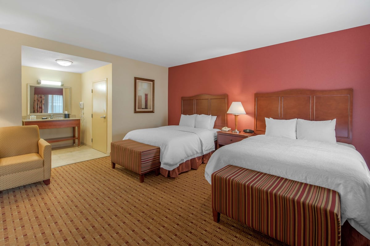 Quarto de hotel com duas camas, área de estar com uma cadeira, uma mesinha e um banquinho.