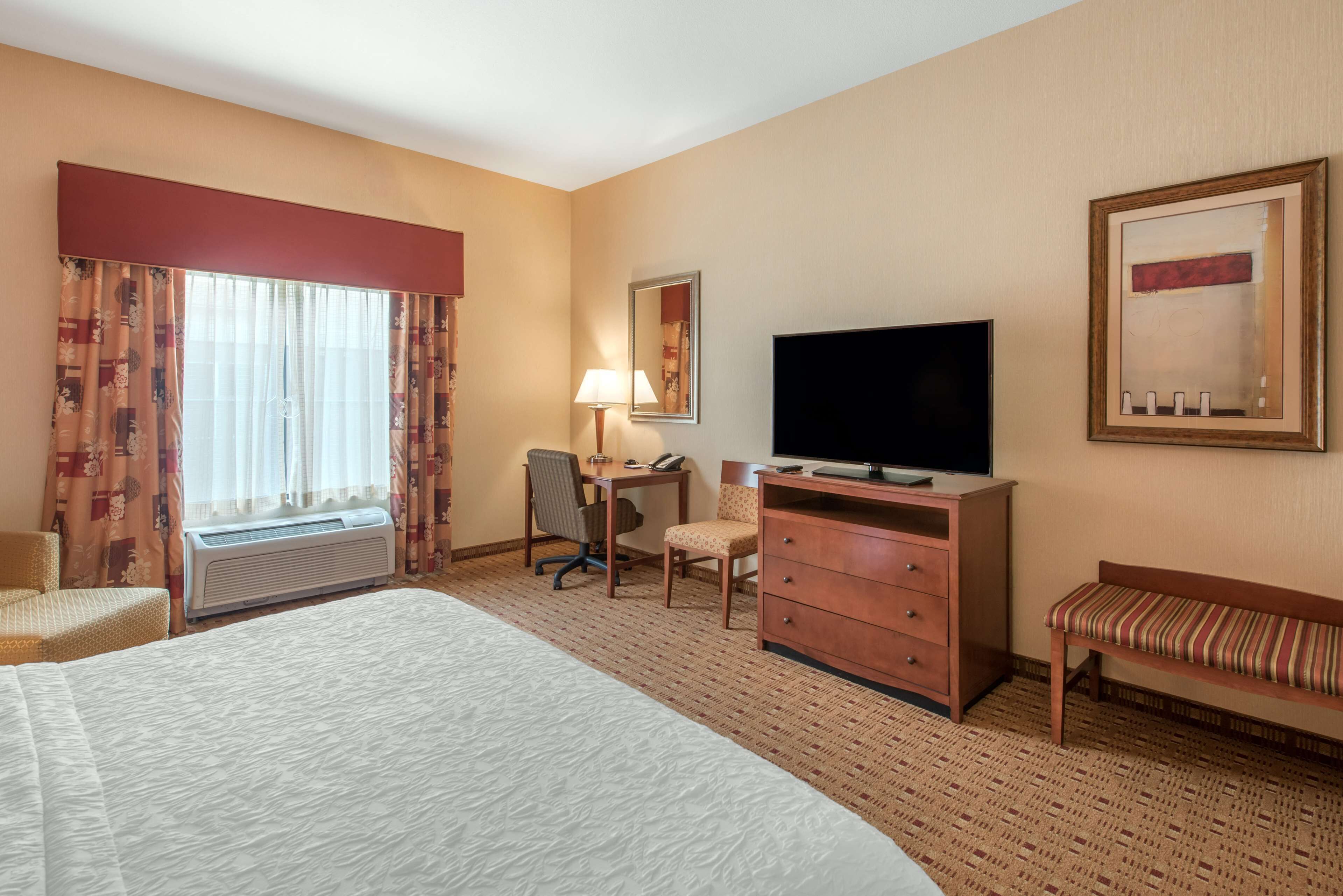 Una camera d'hotel con un letto grande, una scrivania con una sedia, un televisore, una panca e una finestra con tende.