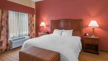 A hotel room with a large bed, two bedside tables with lamps, a window with curtains, and an air conditioning unit.