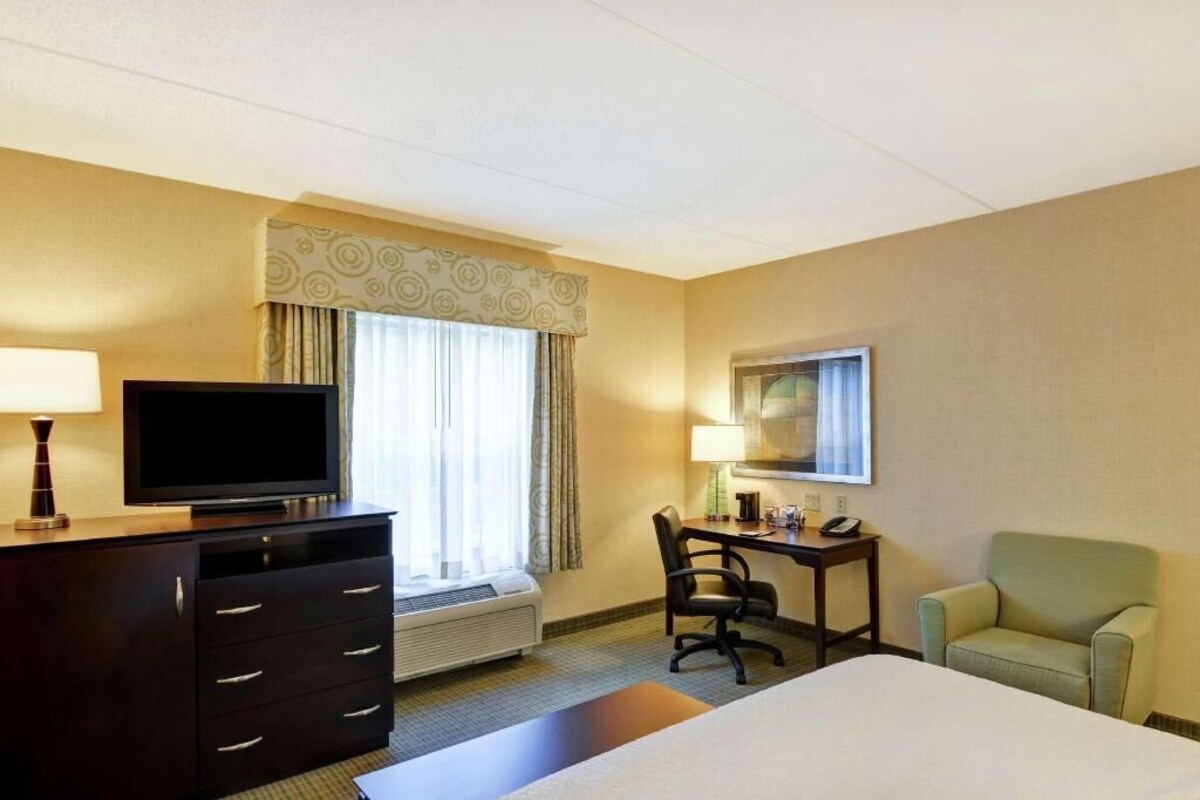 A hotel room with a bed, a desk, a chair, a television, a lamp, and a window with curtains.