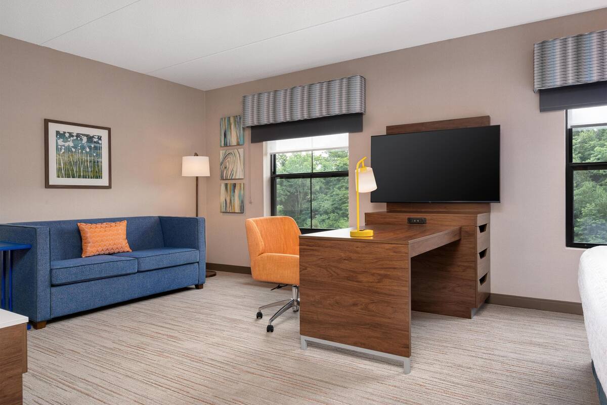 Une chambre d’hôtel moderne avec un canapé bleu, un fauteuil orange, un bureau en bois, un téléviseur à écran plat et des œuvres d’art accrochées aux murs.