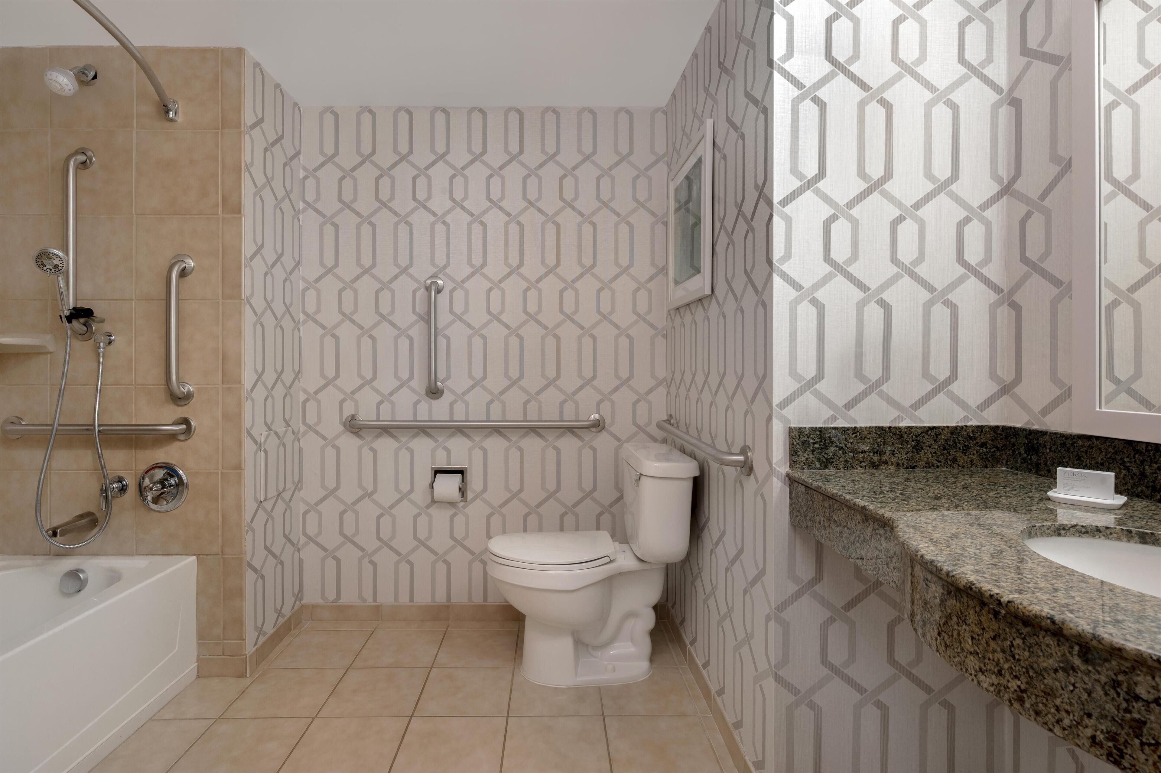 Un baño moderno con inodoro blanco, ducha con barras de apoyo y encimera de granito.