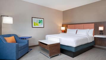 A hotel room with a bed, a blue armchair, a small table, and a nightstand with a lamp.