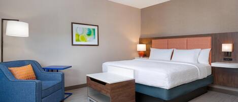 A hotel room with a bed, a blue armchair, a small table, and a nightstand with a lamp.