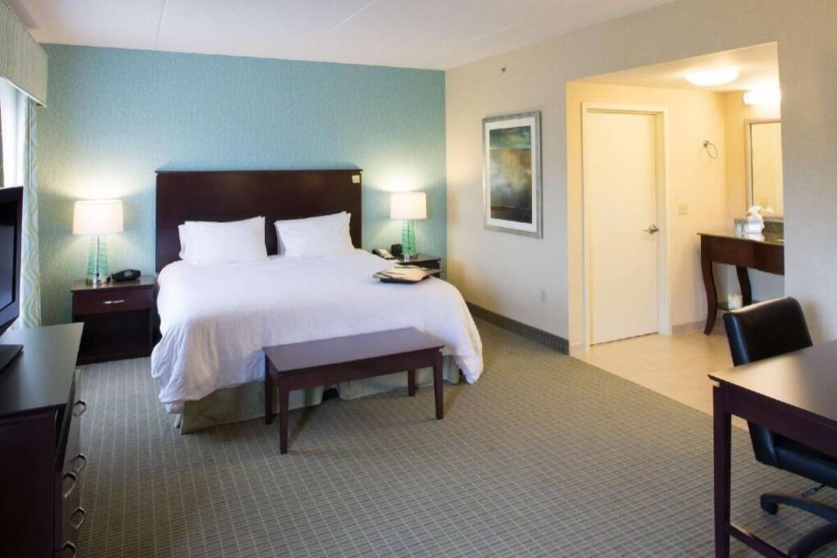 Une chambre d’hôtel comprenant un lit, des tables de chevet, un bureau, une chaise, un banc, un miroir et une porte.