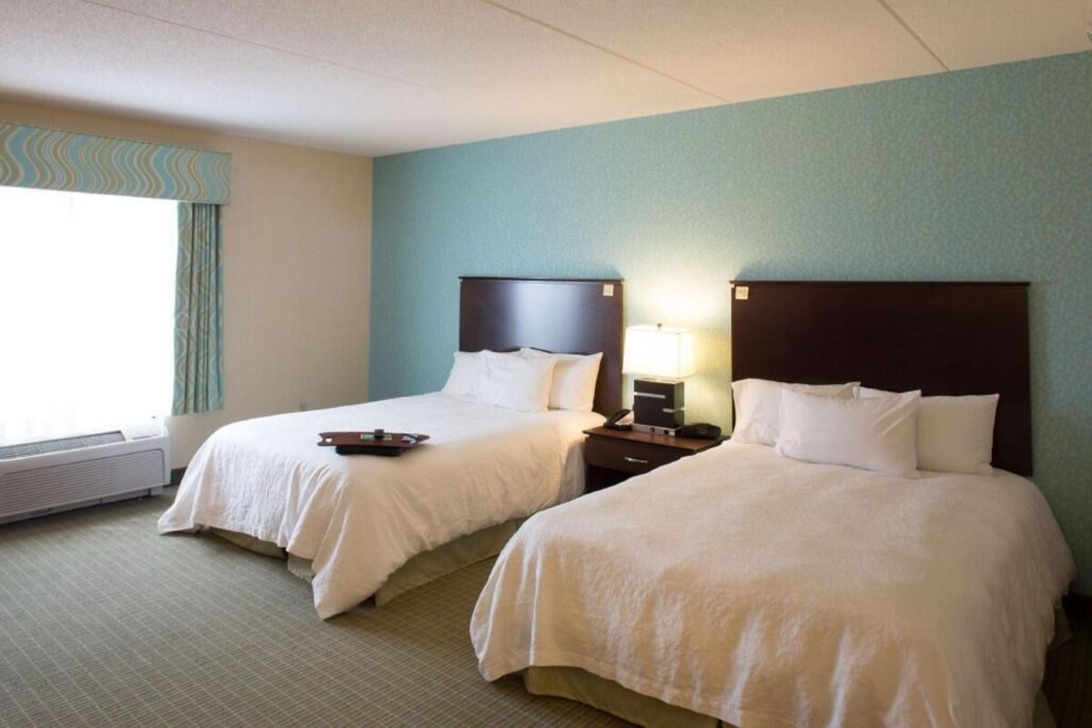 A hotel room with two beds, a window with curtains, a bedside table with a lamp, and a thermostat.