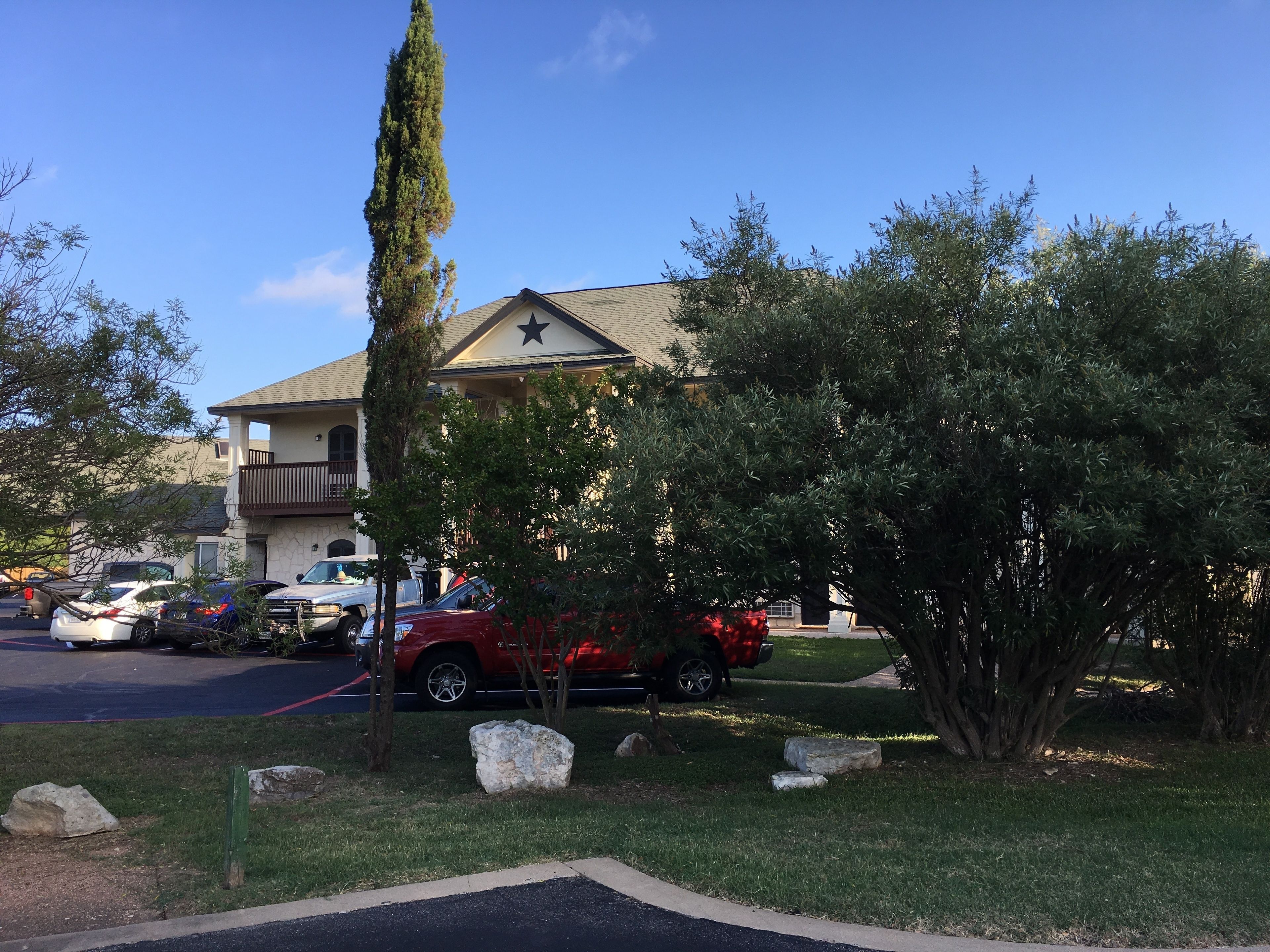 A residential area with a house featuring a balcony and a star on the roof. There are several cars parked in the driveway, and the area is surrounded by trees and rocks.