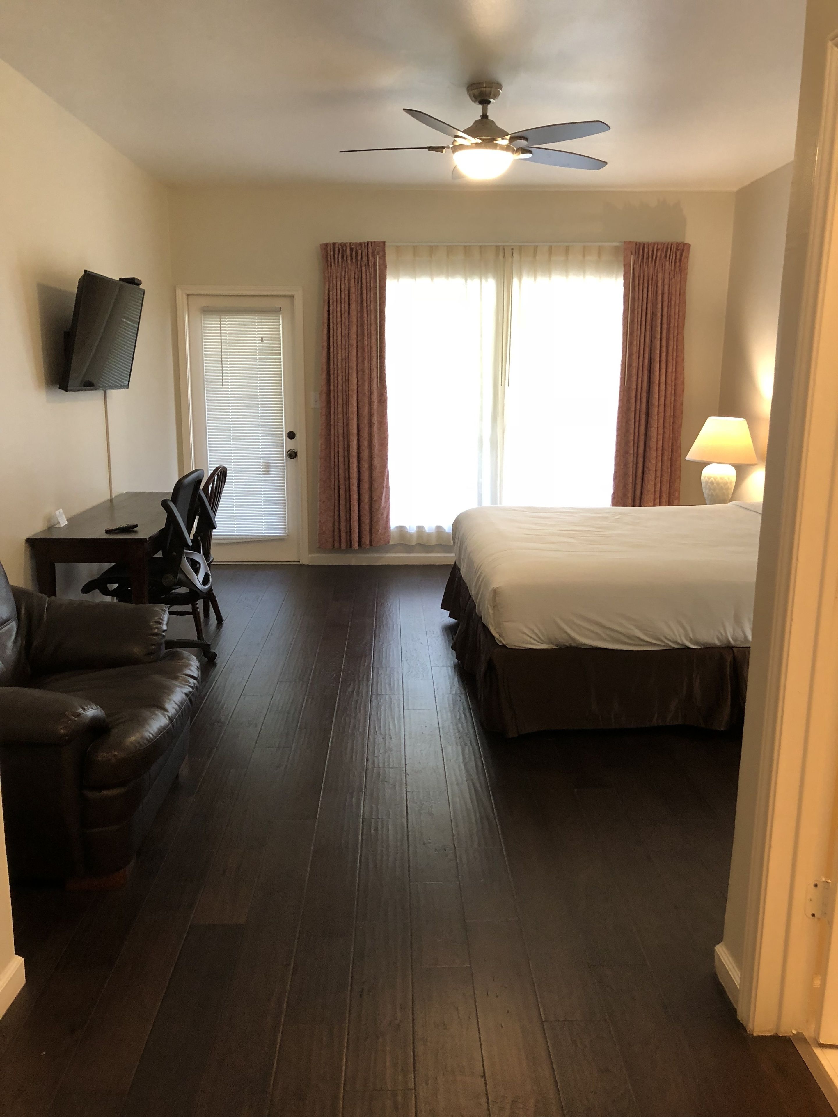 A hotel room with a bed, a desk with a chair, a TV mounted on the wall, and a ceiling fan.