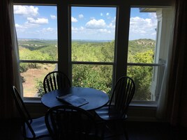 A room with a table and chairs facing a large window with a view of a green landscape and a clear sky.