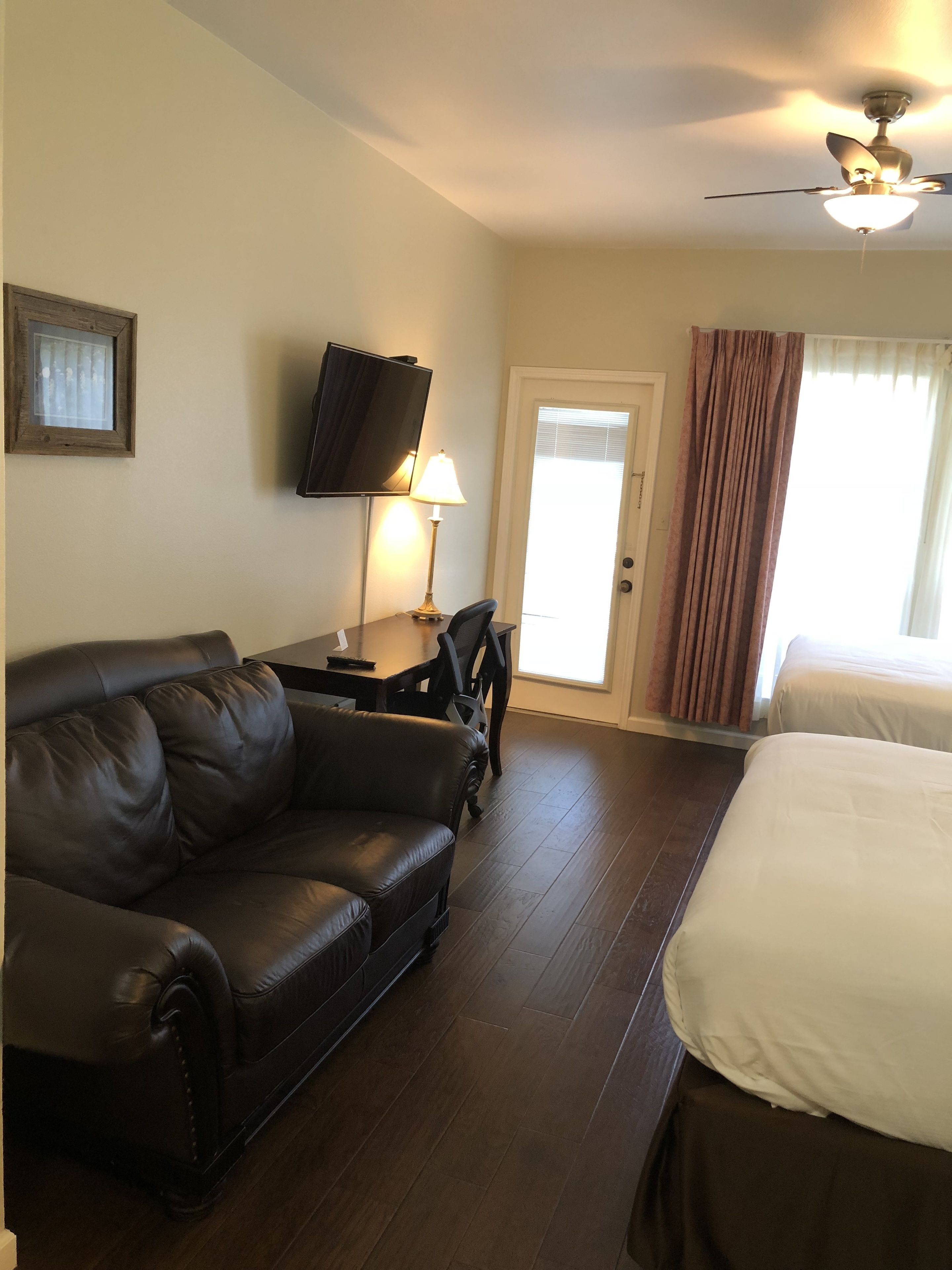 A hotel room with a leather sofa, a bed, a television mounted on the wall, a lamp, and a ceiling fan.