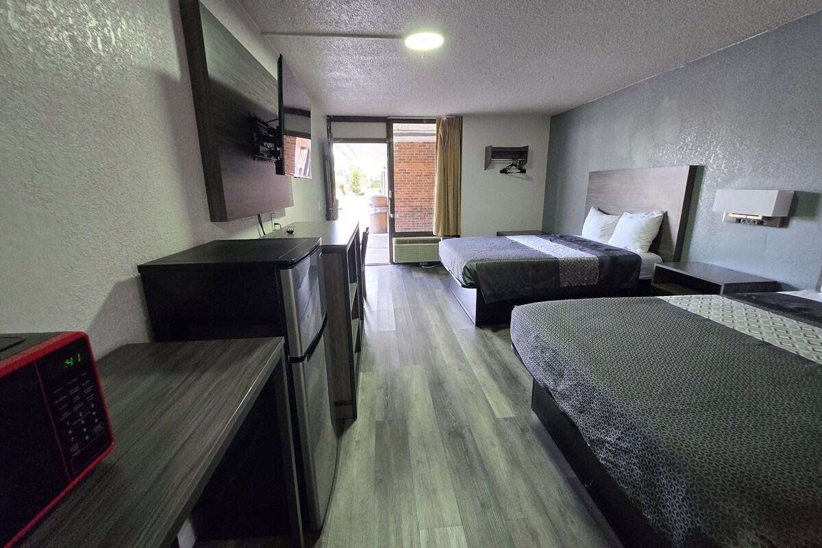 A hotel room with two beds, a desk, a TV, and a kitchenette.