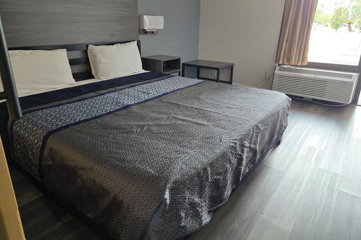 A hotel room with a large bed, a bedside table, a nightstand, a wall-mounted air conditioner, and a window with curtains.