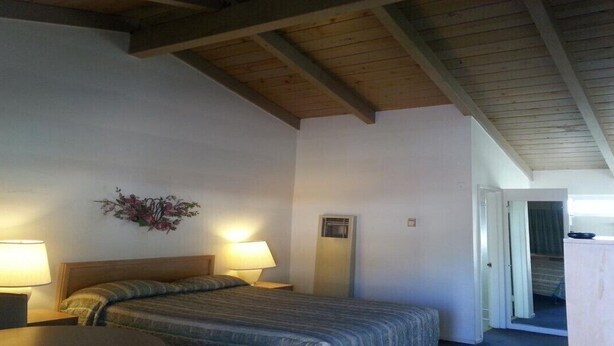A bedroom with a bed, two bedside lamps, a small table, a chair, a wooden ceiling, and a door leading to another room. - Villa Motel (San Luis Obispo)