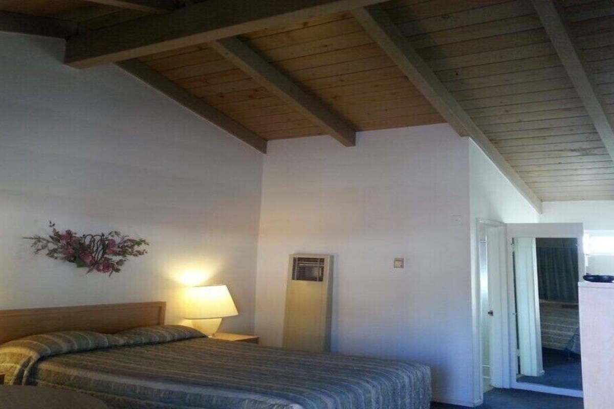 A bedroom with a bed, two bedside lamps, a small table, a chair, a wooden ceiling, and a door leading to another room.