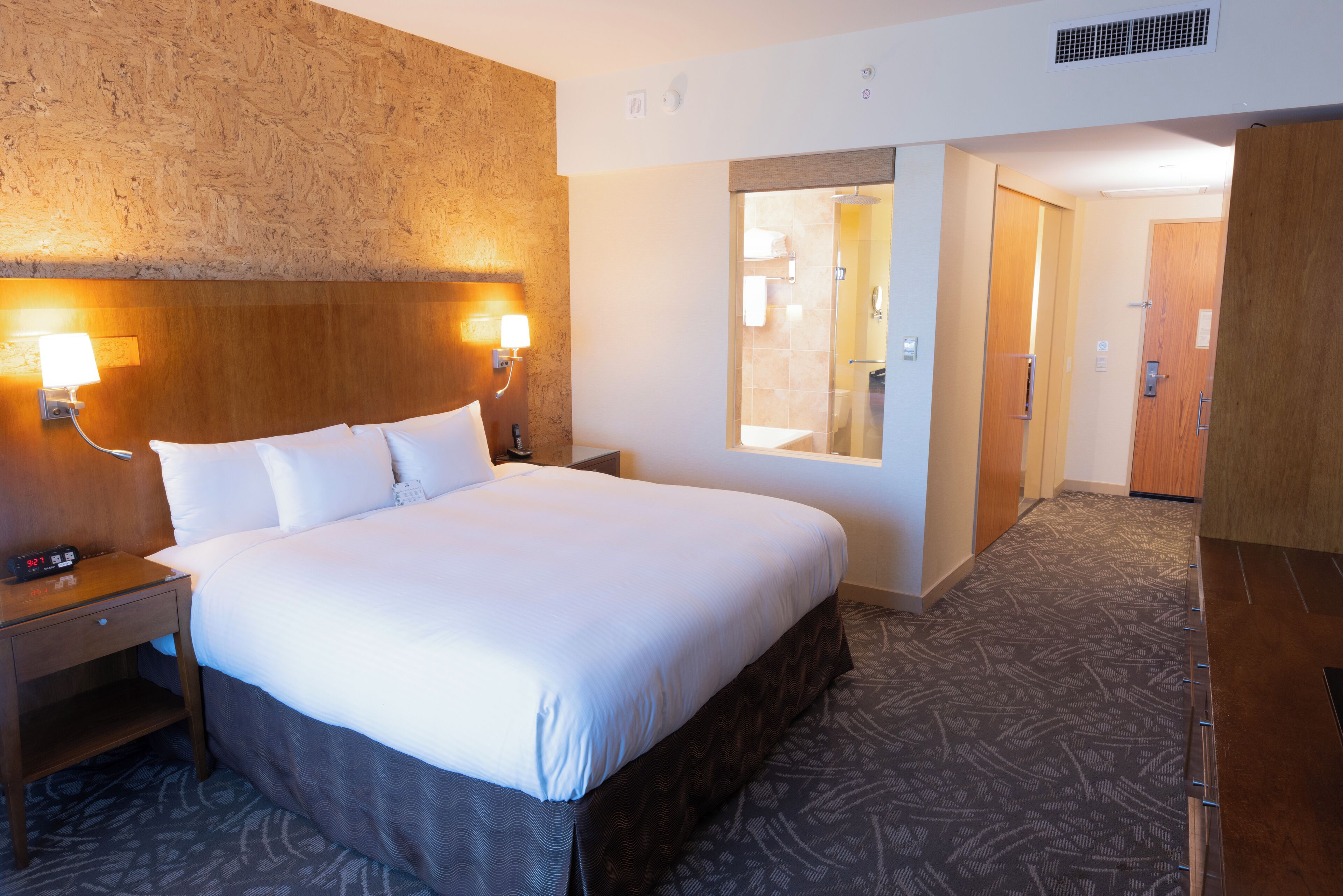 Habitació d'hotel amb un llit gran, abacines, una tauleta de nit, un capçal de llit de fusta, terra amb moqueta i un bany visible a través d'una porta oberta.