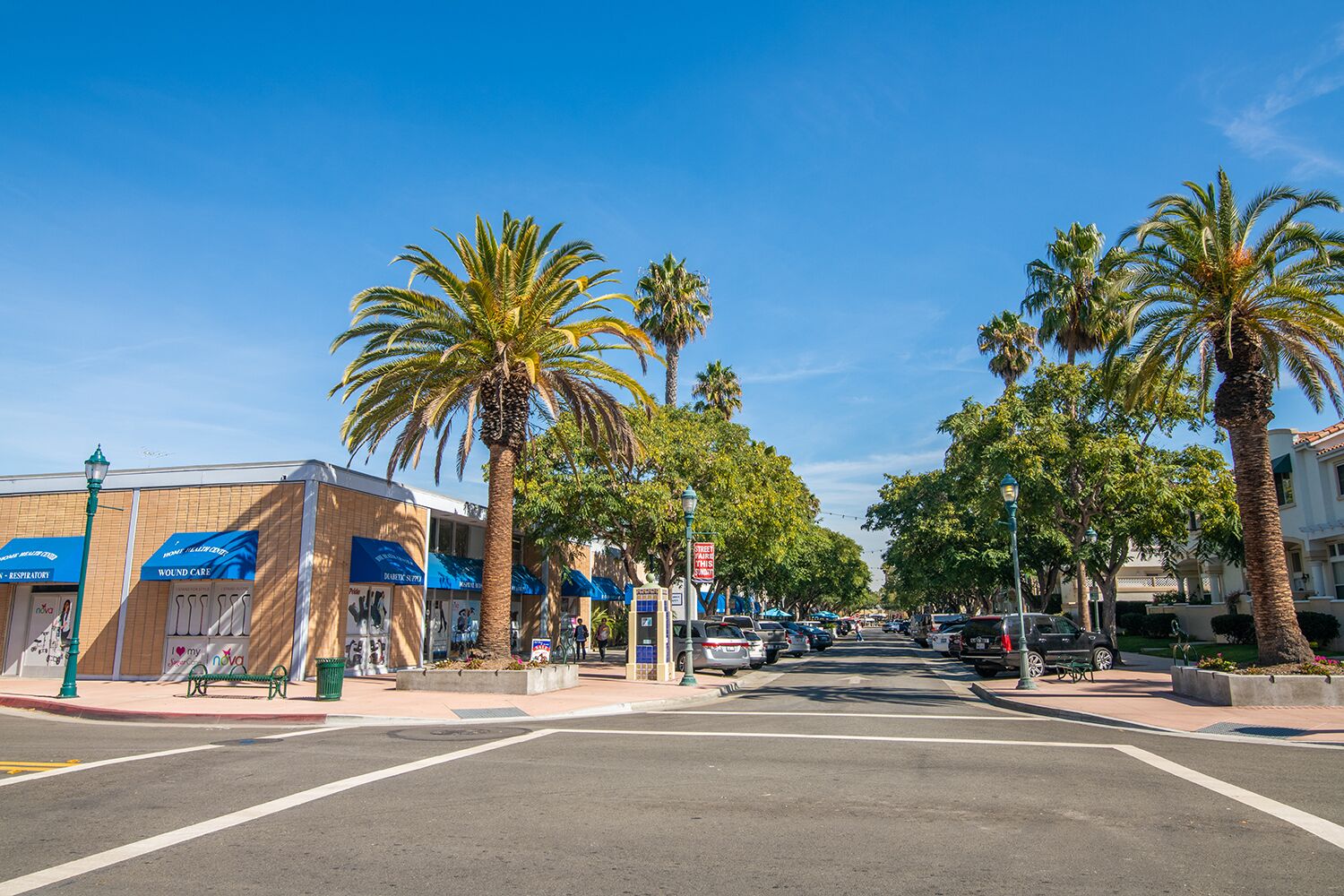 A street with palm trees, a sidewalk, and buildings with blue awnings.