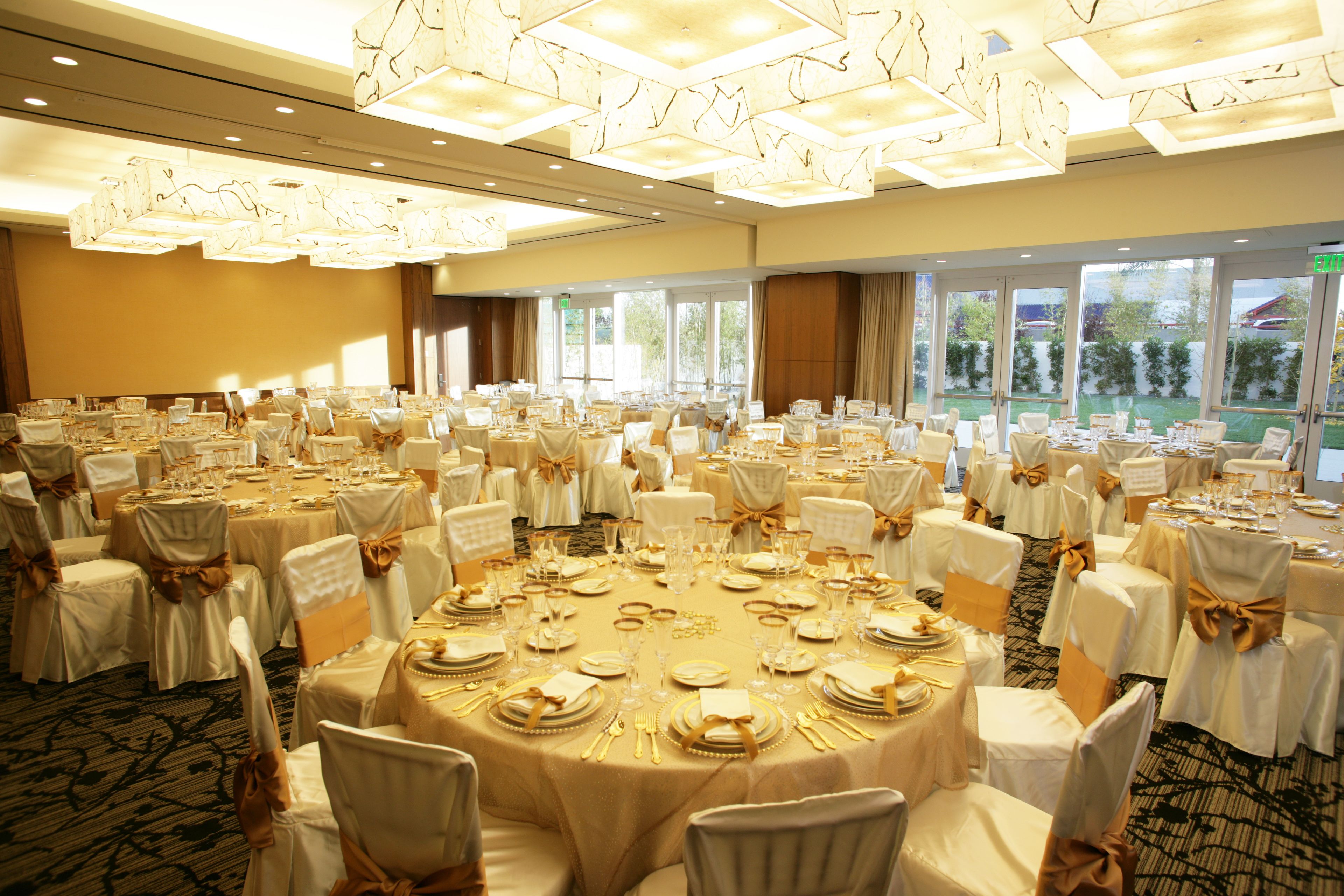 Un salón de banquetes con mesas redondas dispuestas para comer, manteles blancos y sillas con cintas doradas.