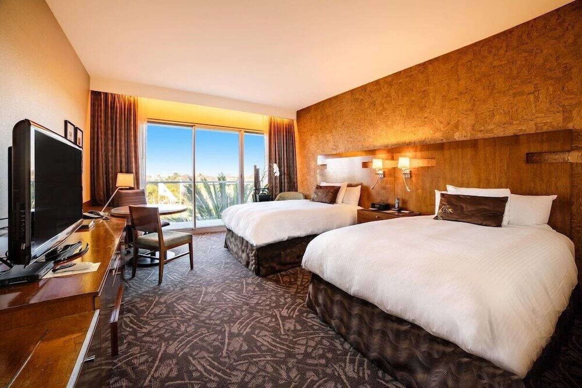 A hotel room with two beds, a desk with a TV, and a large window with a view.