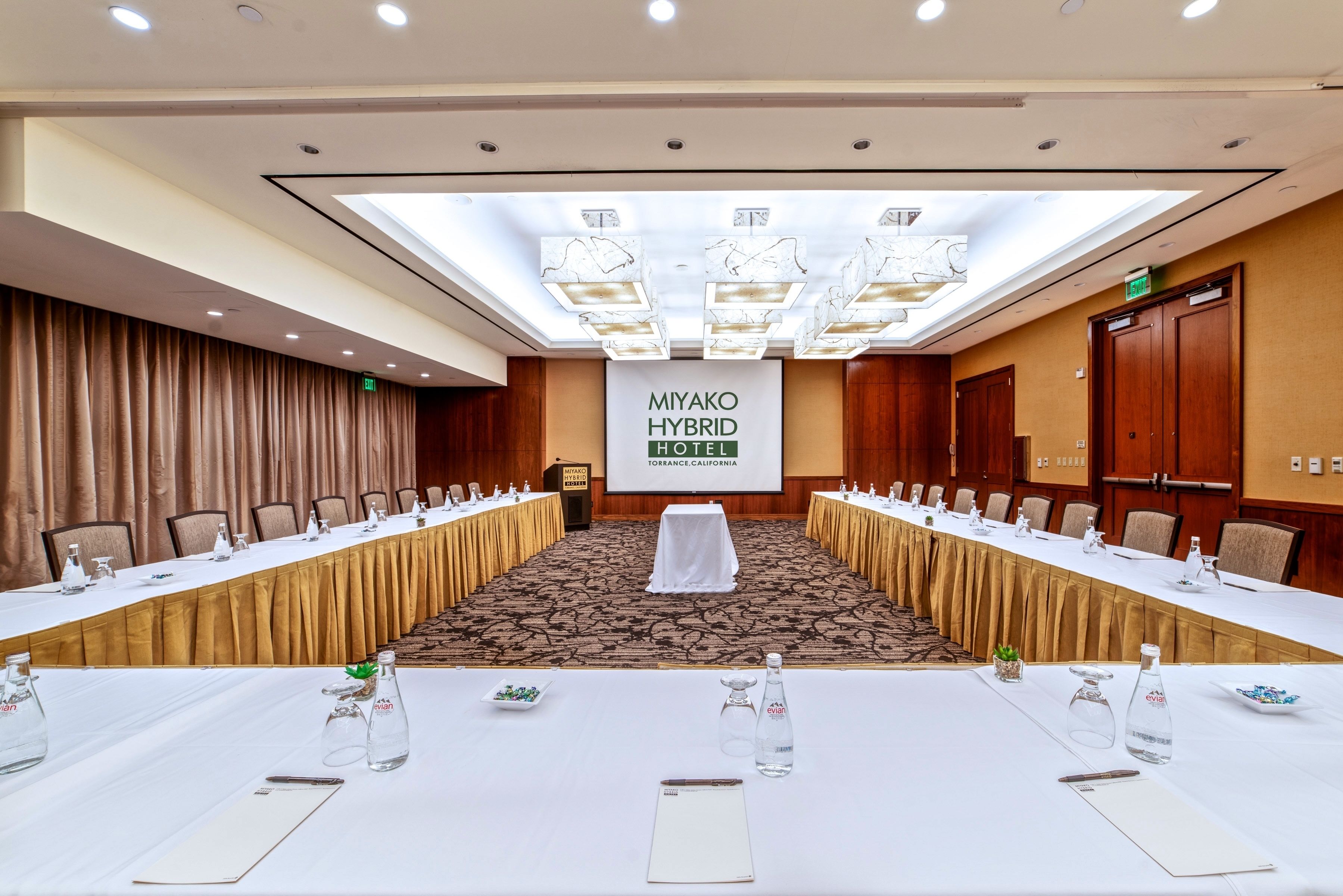 Ett konferensrum med ett långt bord, stolar och en talarstol. En banderoll visar texten ”MIYAKO HYBRID HOTEL” och ”TORRANCE, KALIFORNIEN”.