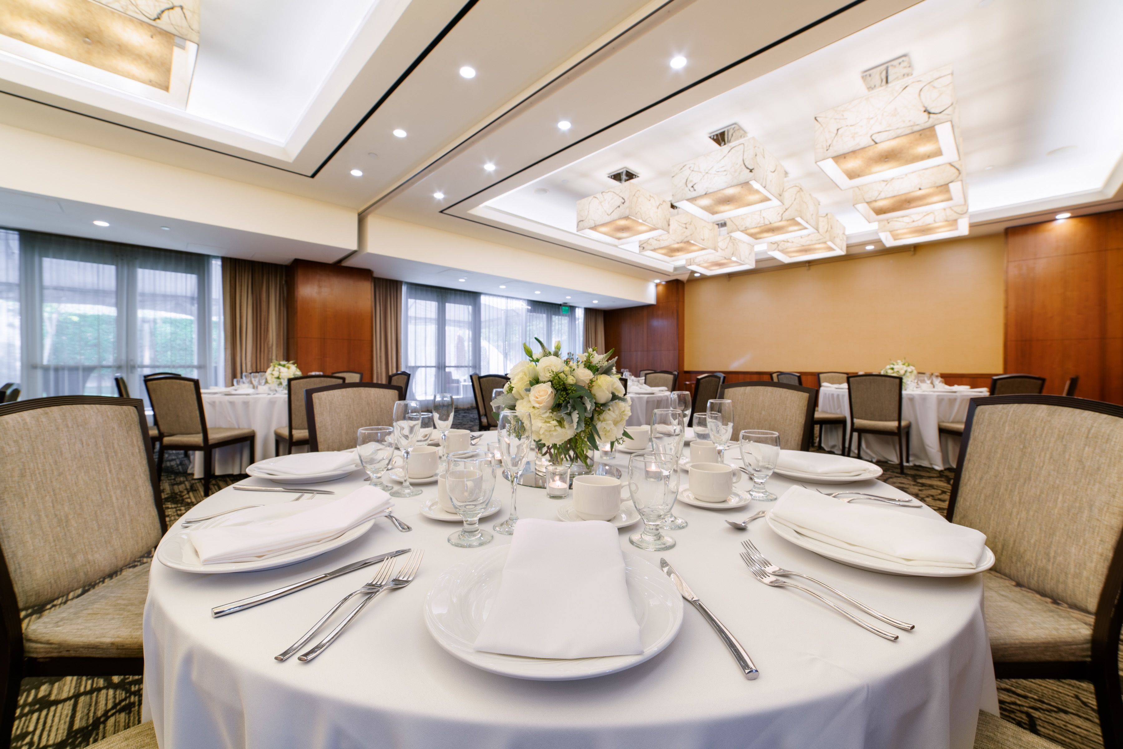 A banquet hall with round tables set for dining, featuring white tablecloths, plates, cutlery, and floral centerpieces.