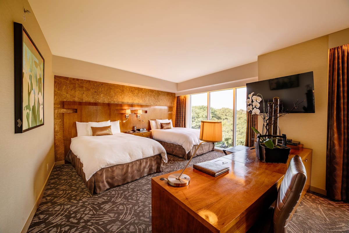 A hotel room with two beds, a wooden desk, a television, and a large window with curtains.