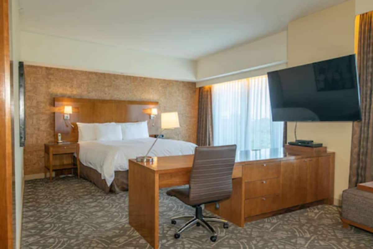 A hotel room with a bed, desk, chair, TV, and a large window with curtains.