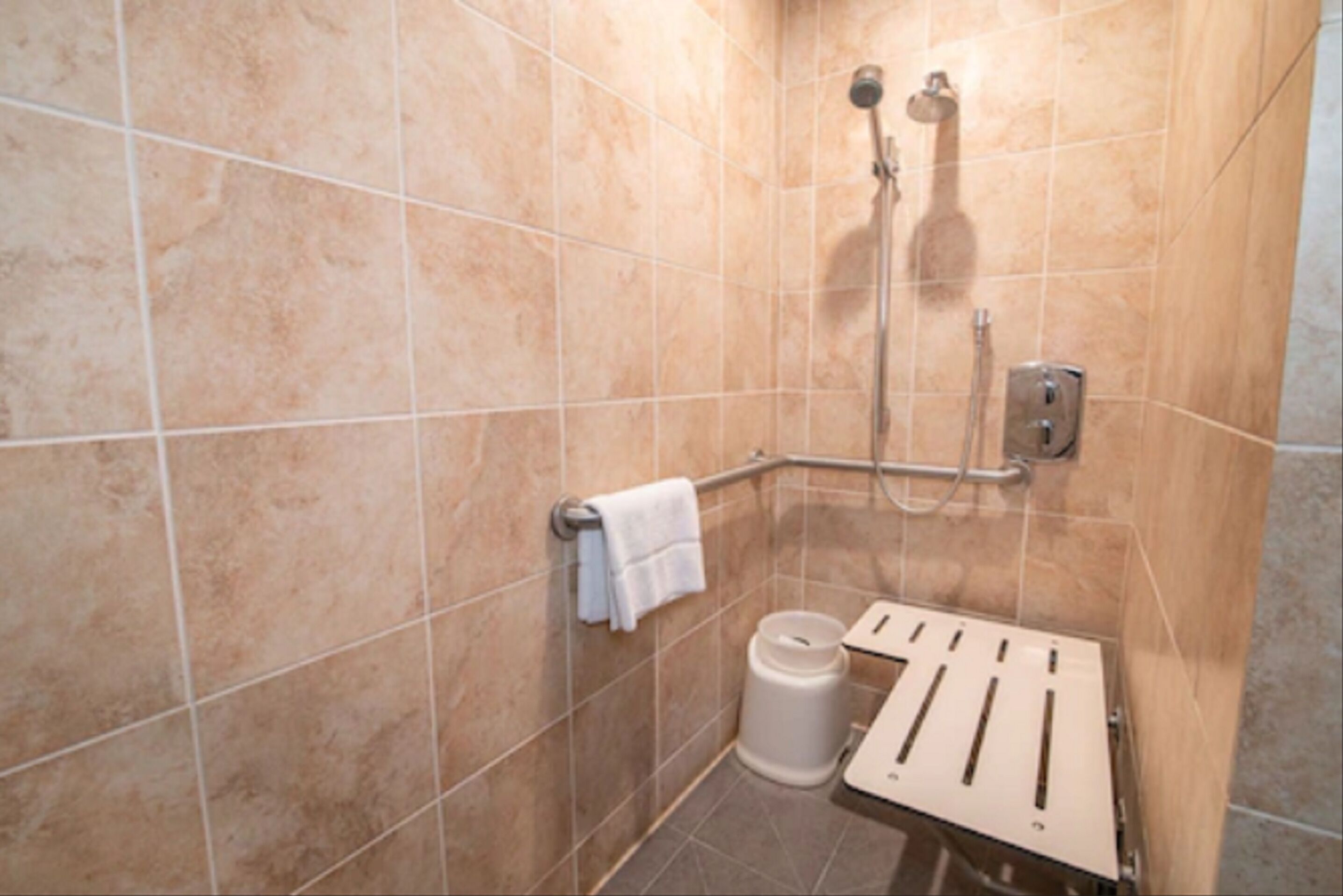 A bathroom with beige tiles, a shower with a handheld showerhead, a grab bar, a towel rack, and a toilet paper holder.
