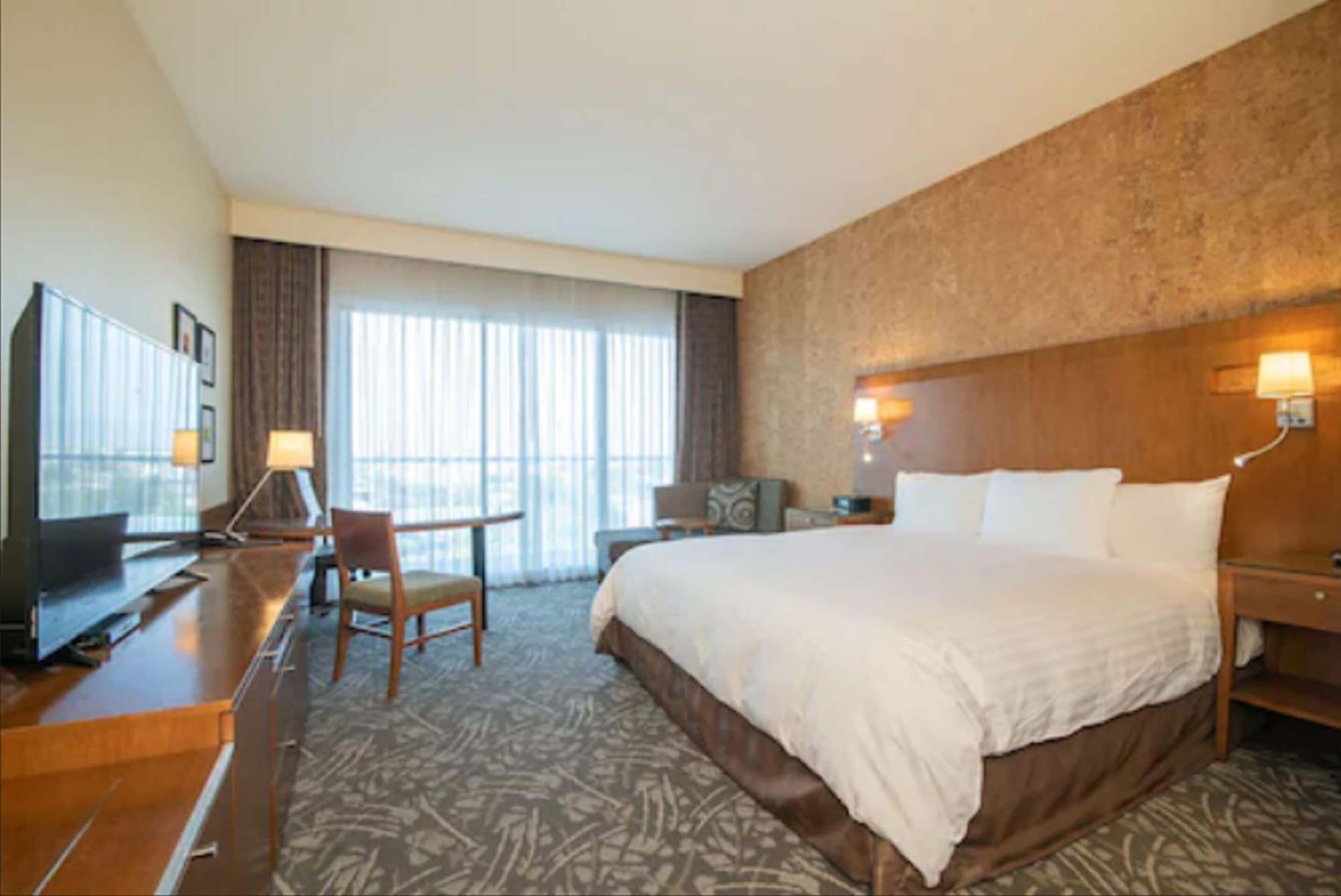 A hotel room with a large bed, a desk with a chair, a television, and a window with curtains.
