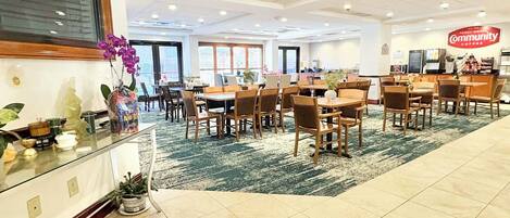 Un amplio vestíbulo de hotel con varias mesas y sillas de comedor, una zona de recepción con mostrador y un letrero de cafetería.