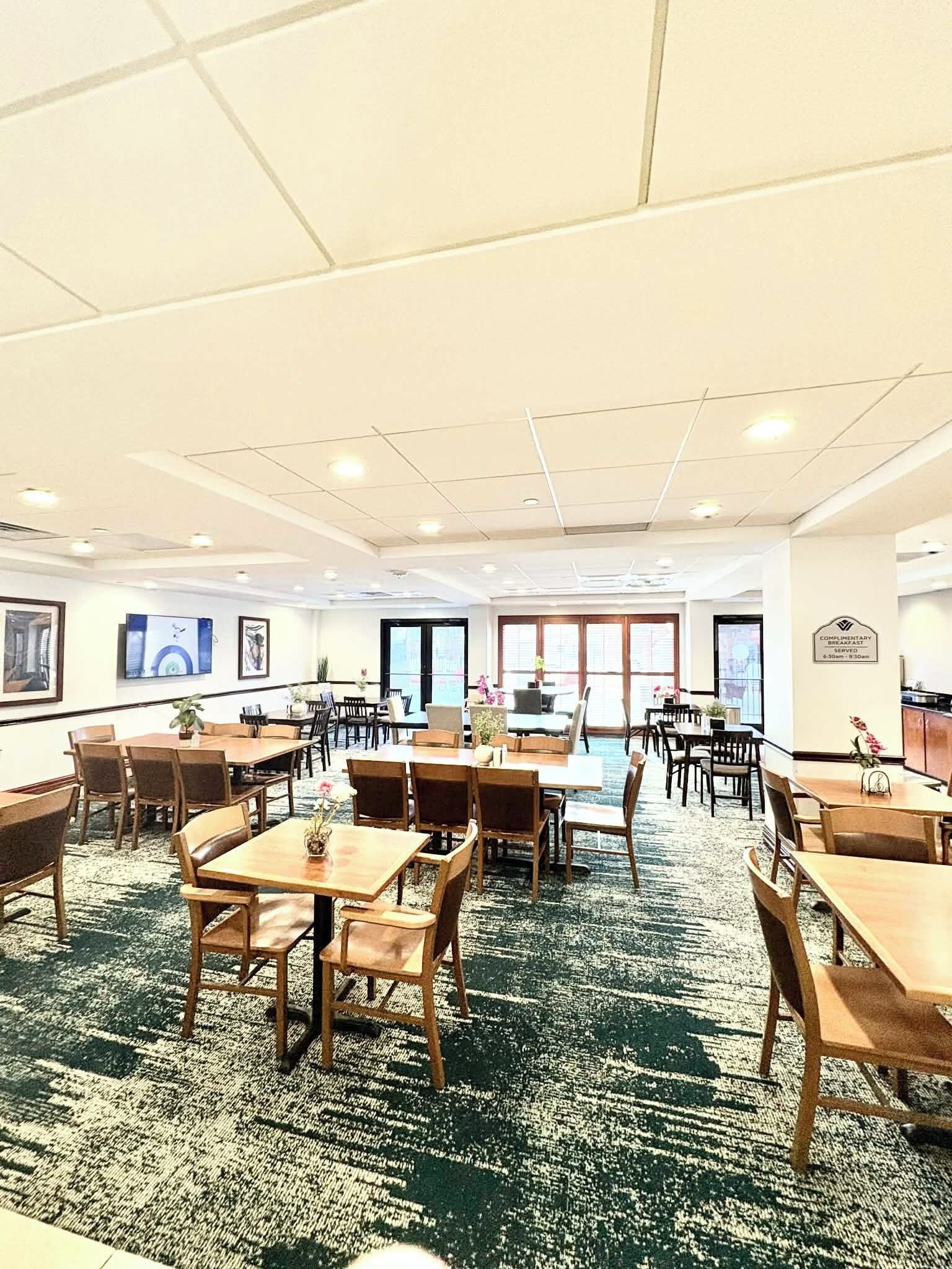 A spacious hotel dining area with multiple tables and chairs, large windows, and a carpeted floor.