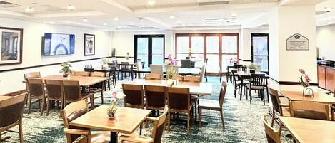 A spacious hotel dining area with multiple tables and chairs, large windows, and a carpeted floor.