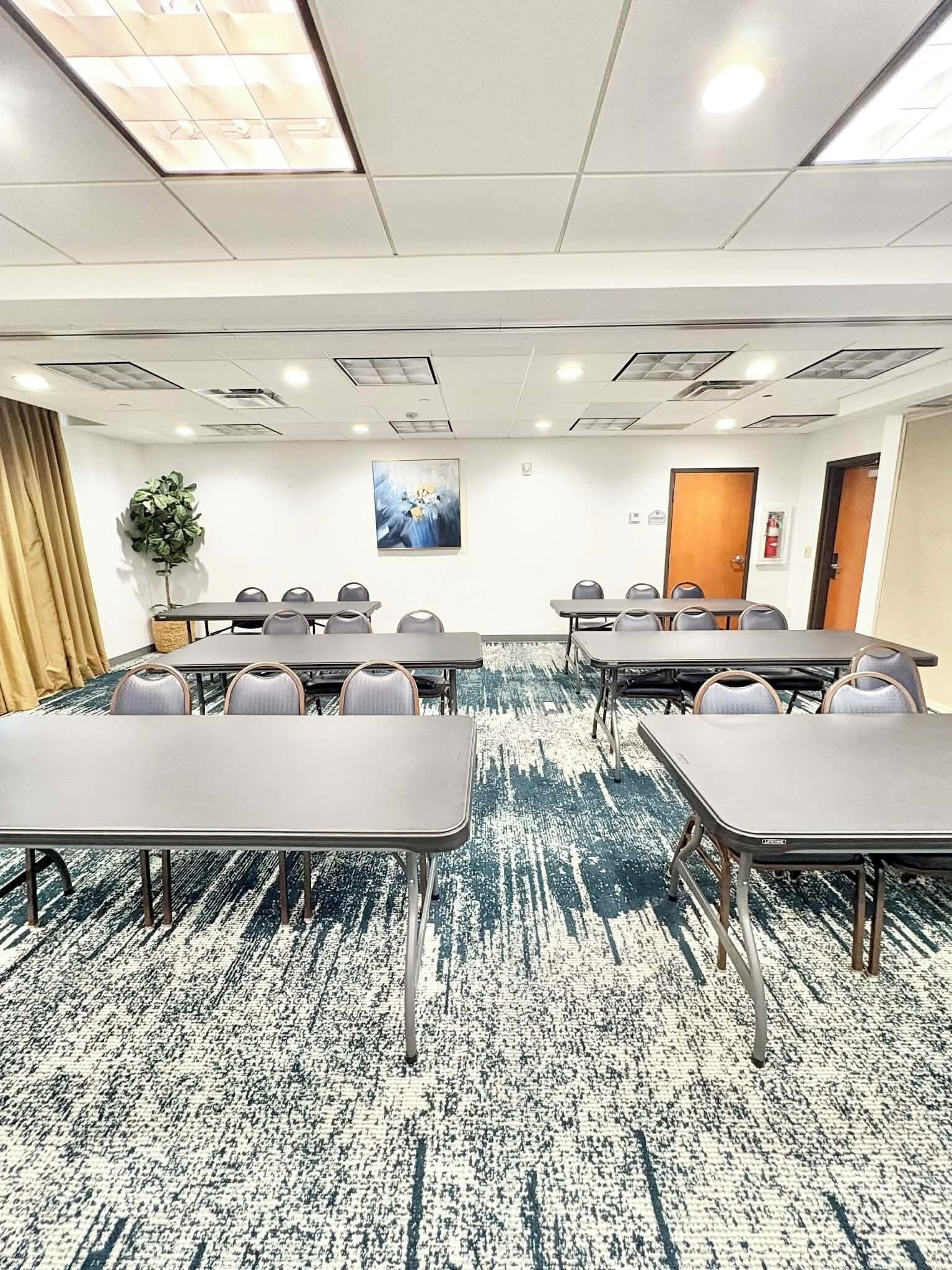A conference room with multiple tables and chairs, a patterned carpet, ceiling lights, and a door.