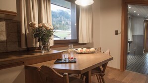Un coin repas avec une table en bois, des chaises, un vase de fleurs, un panier de fruits et une vue sur un paysage montagneux par la fenêtre.