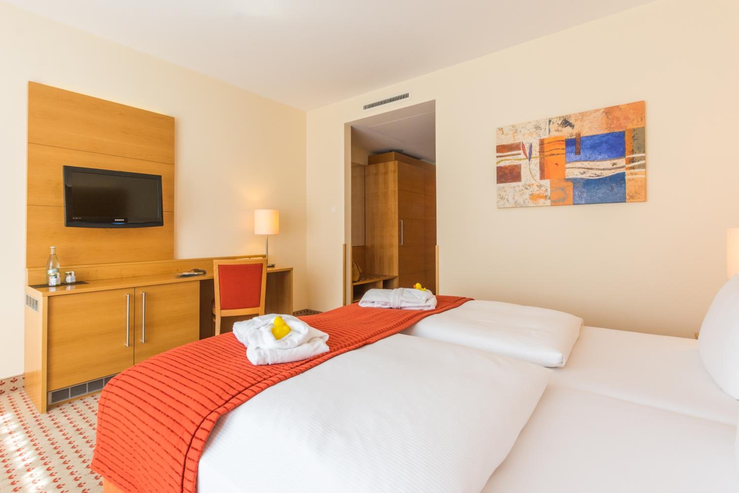 Una habitación de hotel con cama, escritorio, televisor, un cuadro y alfombra roja.