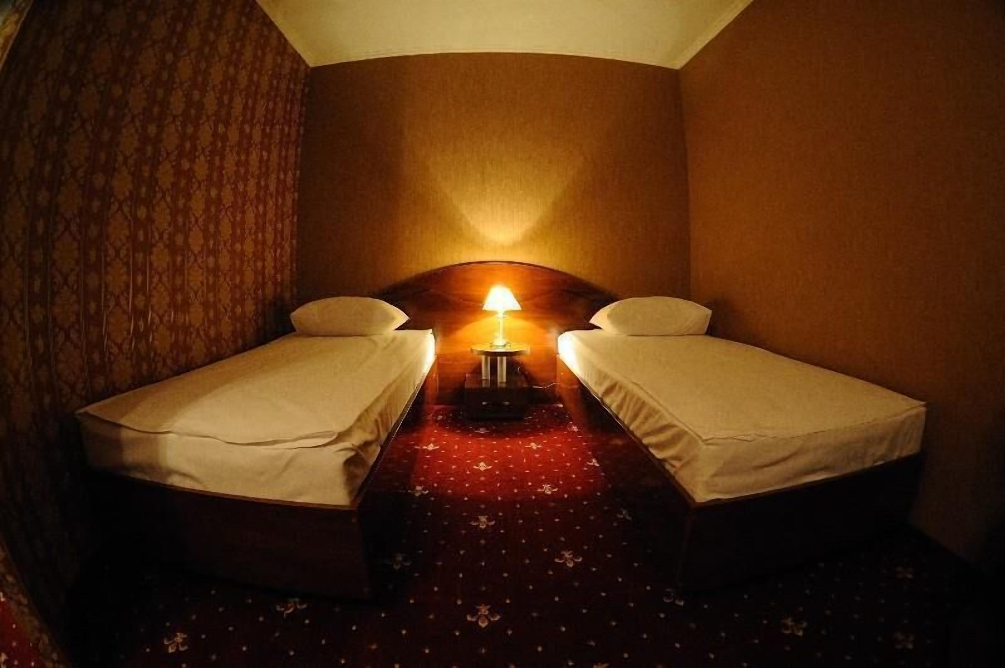 Una habitación pequeña y tenuemente iluminada con dos camas individuales, una mesita con una lámpara y una alfombra estampada.