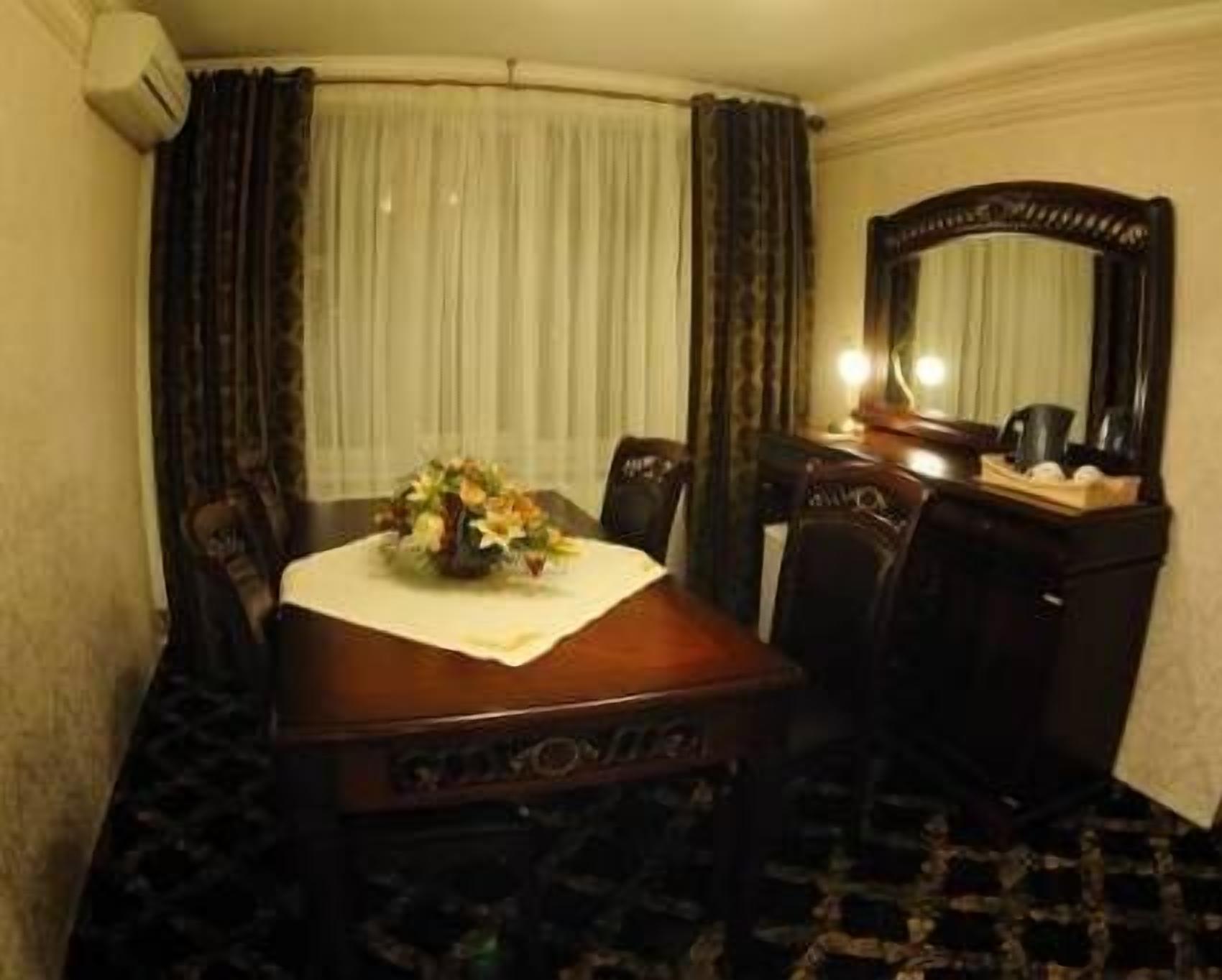 Habitación de hotel con mesa de comedor de madera, sillas, un jarrón de flores, un espejo y una ventana con cortinas.