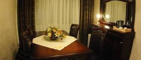 Habitación de hotel con mesa de comedor de madera, sillas, un jarrón de flores, un espejo y una ventana con cortinas.