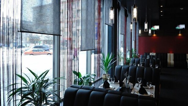 A restaurant with a modern interior, featuring a long dining table with black upholstered chairs, potted plants, and a view of the street through large windows. - Mir Hotel In Rovno (Rivne)