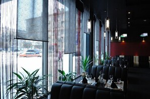 A restaurant with a modern interior, featuring a long dining table with black upholstered chairs, potted plants, and a view of the street through large windows. - Mir Hotel In Rovno (Rivne)
