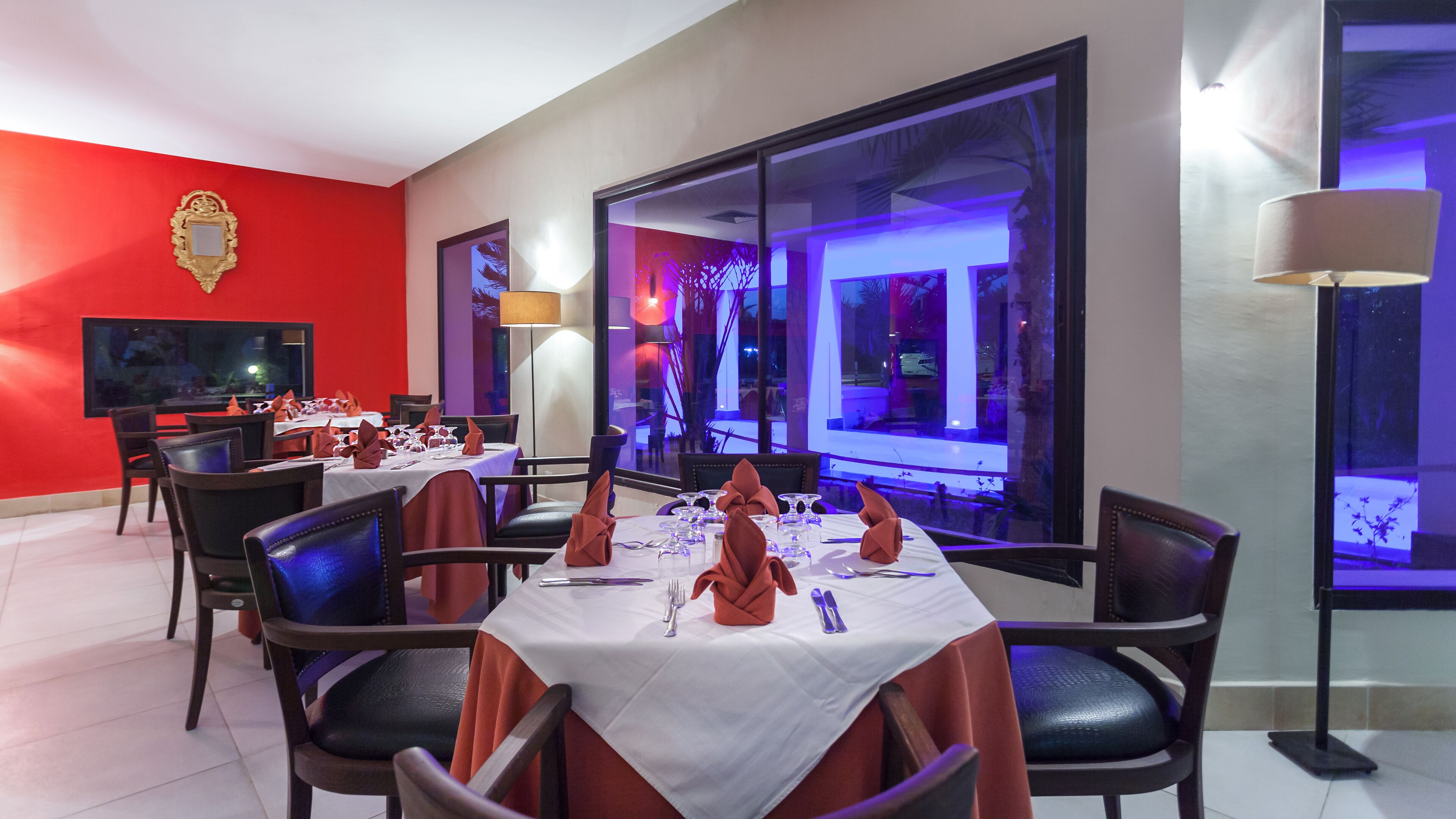 Comedor con mesas dispuestas para una comida, sillas, un televisor y un amplio ventanal con vistas al exterior.