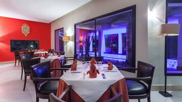 Comedor con mesas dispuestas para una comida, sillas, un televisor y un amplio ventanal con vistas al exterior.